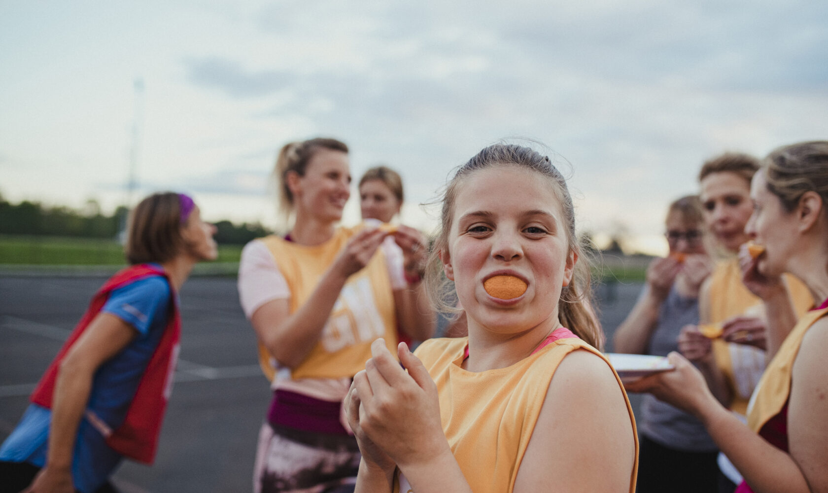 A girl places an orange slice in her mouth and smiles at the camera after a netball game. Her teammates can be seen behind her.