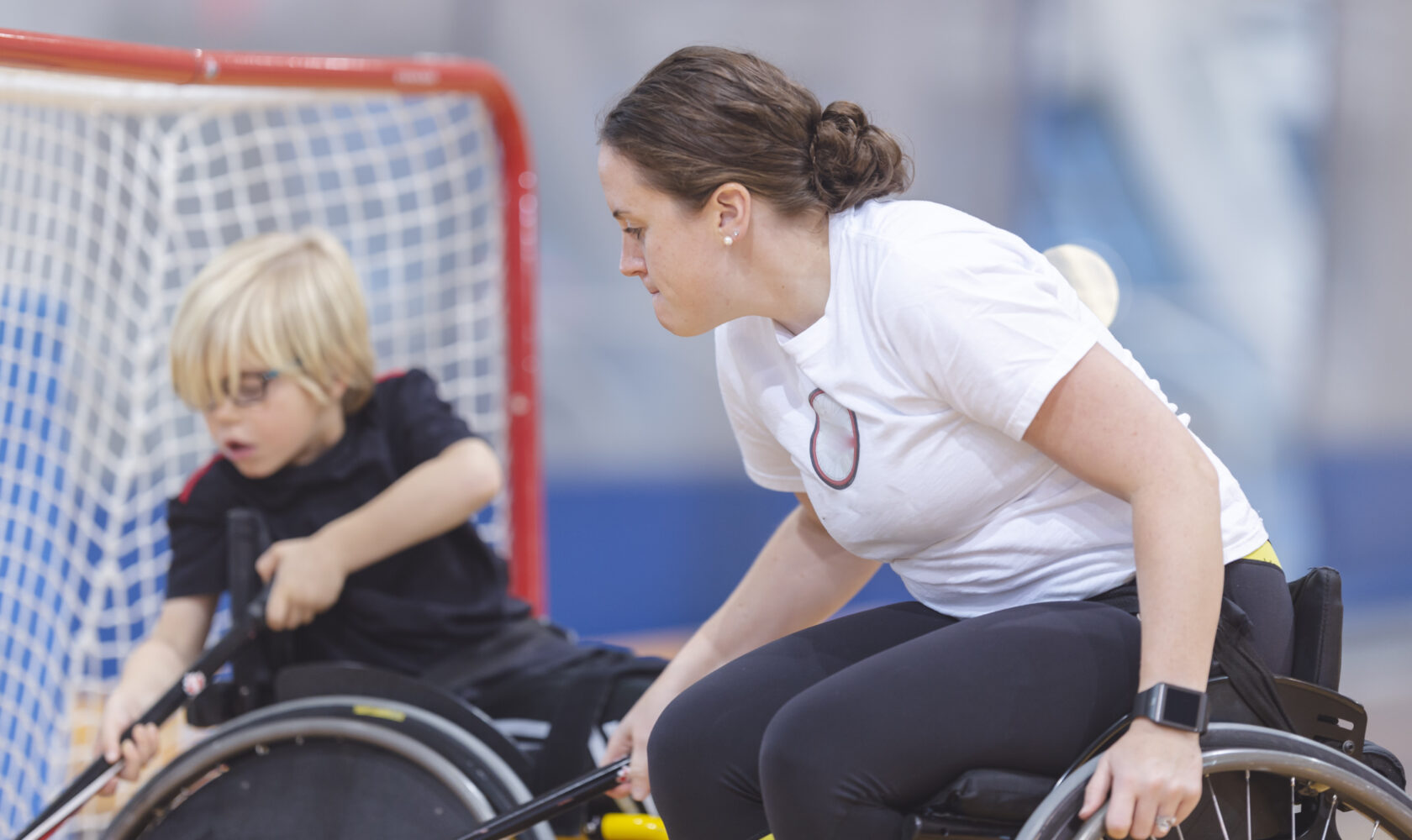 A female coach in a wheelchair wearing a white t-shirt plays lacrosse with a young boy also in a wheelchair.